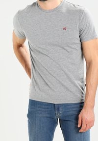 Šedé bavlněné tričko s krátkými rukávy a kulatým výstřihem. Na levé straně hrudi je malá červenobílá logo. Kombinováno s modrými džínami.