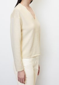 Beiger Strickpullover mit V-Ausschnitt und langen Ärmeln. Hat eine strukturierte Stoffoberfläche und einen geraden Schnitt am Saum, kombiniert mit weißen Hosen.