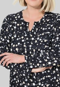 Chemise noire avec des motifs circulaires blancs et crème, à manches longues et un petit décolleté en trou de serrure. Le tissu semble doux et texturé.