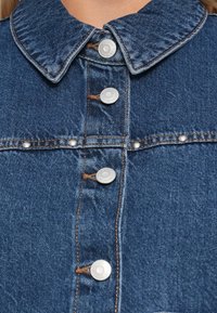 Denim košulja s klasičnim ovratom, pet srebrnih dugmadi na pritisak, plava tkanina s vidljivim šavovima i blago teksturiranom površinom.