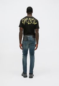 Homem de pé, de costas, vestindo uma t-shirt preta com o texto amarelo "Diesel" e jeans azuis desbotados, com ténis pretos, em fundo branco.