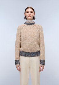 Gruby sweter dziany w beżowym kolorze z teksturowanymi ściegami. Rękawy i dół wykończone ribbitem w niebiesko-brązowo-kremowym wzorze. Modelka nosi kremowe spodnie.