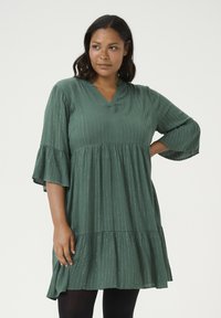 Vestido verde de longitud hasta la rodilla, hecho de una tela texturizada con rayas verticales, escote en V y mangas acampanadas. Detalle de dobladillo de volantes en capas.