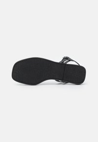 Svart platt sandal med en texturerad gummisole med ett geometriskt mönster, justerbart rem och minimalistisk design.