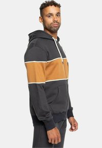 Sudadera con capucha en gris oscuro con rayas horizontales en color beige y blanco. Cuenta con un cierre frontal y bolsillos; está hecha de un tejido suave y texturizado.
