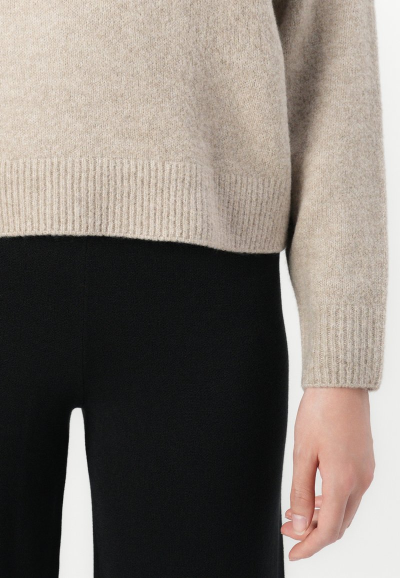 Beige Strickpullover mit geripptem Saum und Bündchen, getragen mit schwarzen eng anliegenden Hosen, das rechte Arm und die Hand entspannt an der Seite zeigend.