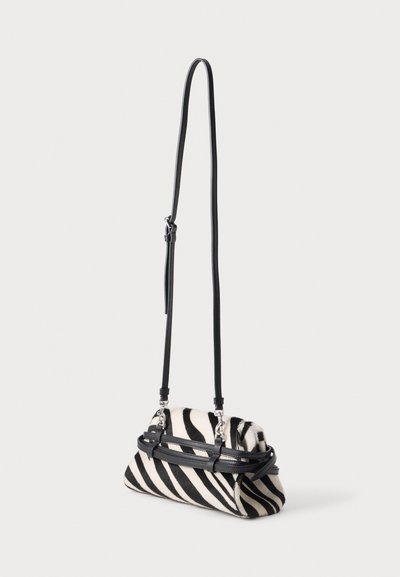 Petit sac à bandoulière avec des rayures zébrées noires et blanches et une longue sangle en cuir noire réglable suspendue sur un fond clair.