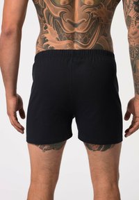 Boxer cortos de algodón negros con una cinturilla elástica, vista desde atrás, mostrando tatuajes intrincados en las piernas y la parte superior de la espalda del modelo.