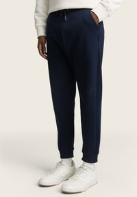 Pantalones jogger azul marino con puños elásticos y cintura con cordón, con bolsillos laterales. Combinados con zapatillas blancas y calcetines claros.