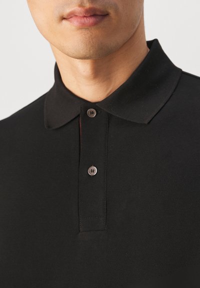 Sort polo skjorte med en klassisk krave, to knap-lukning og struktureret stof. Nærbillede der fremhæver detaljerne i beklædningen.