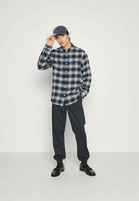 Camisa de flanela xadrez em azul marinho e branco, com frente abotoada, combinada com calças escuras e sapatos pretos. Um boné azul marinho complementa o conjunto.