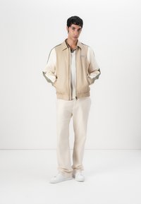 Beige Bomberjacke mit farbblockierten Ärmeln in Weiß und Oliv. Reißverschluss, elastische Bündchen, getragen mit cremefarbenen Hosen und weißen Sneakers.