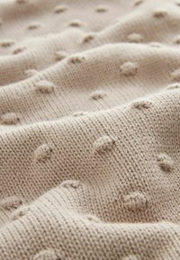 Tessuto lavorato a maglia beige con motivi a punti in rilievo. Morbida texture, spessore medio e una finitura liscia con dettagli di cucitura visibili.