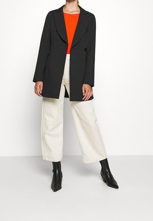 Personne portant un long blazer noir à fines rayures, un top orange vif, un pantalon large écru et des bottines noires à talons pointus.