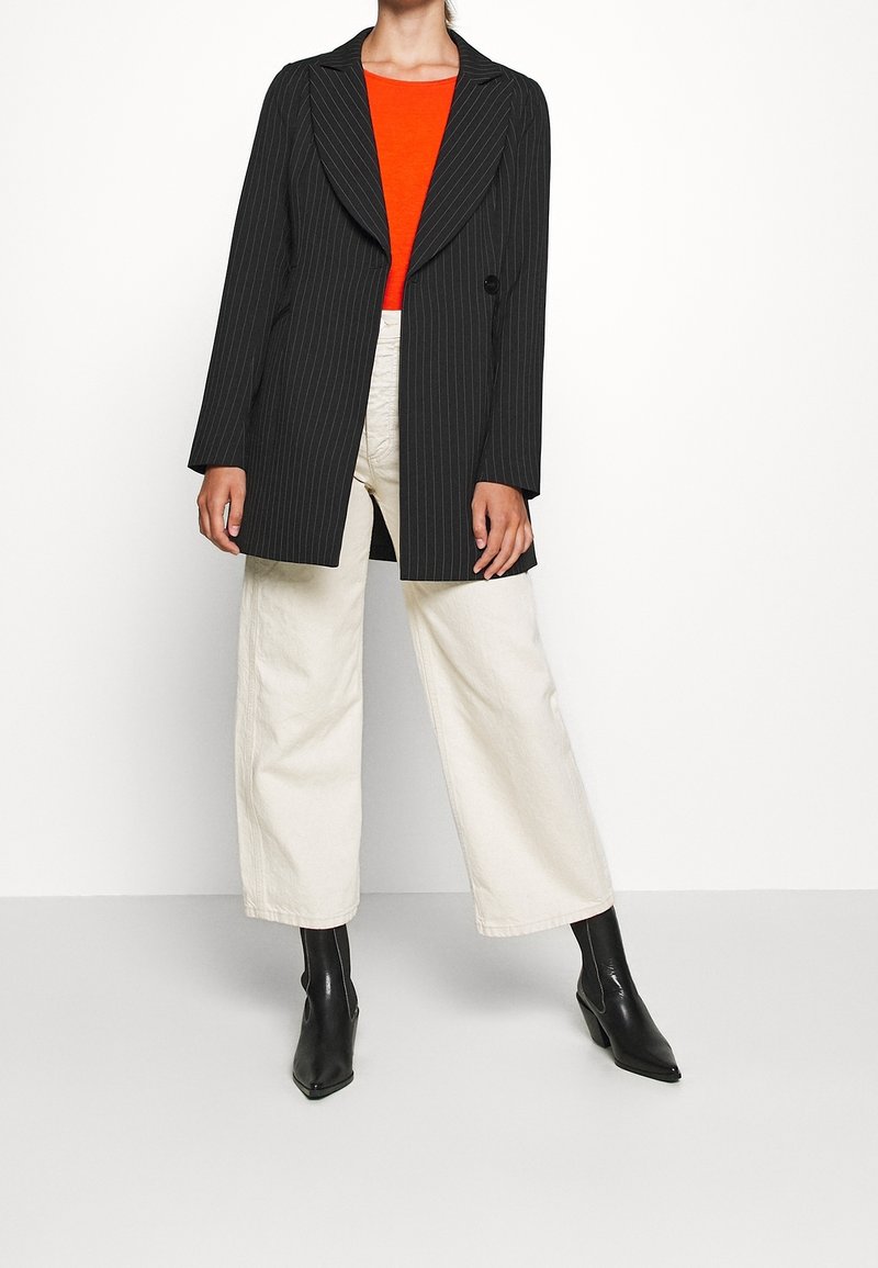 Personne portant un long blazer noir à fines rayures, un top orange vif, un pantalon large écru et des bottines noires à talons pointus.