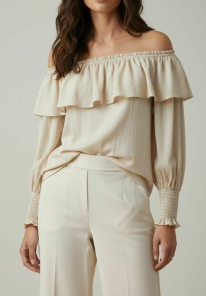 Femme portant un chemisier beige à épaules dénudées avec un col à volants et des manches longues froncées, associé à un pantalon beige taille haute.