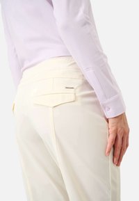 Kobieta w kremowych spodniach z klapką na tylnej kieszeni i jasnoliliowej bluzce z długim rękawem, stojąca z ręką swobodnie opuszczoną.