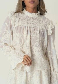 Weiße bestickte Bluse mit gerafftem Ausschnitt und Bündchen, langen Ärmeln, floralen Mustern und einem halbtransparenten Stoff. Mit gewellten Abschlüssen.