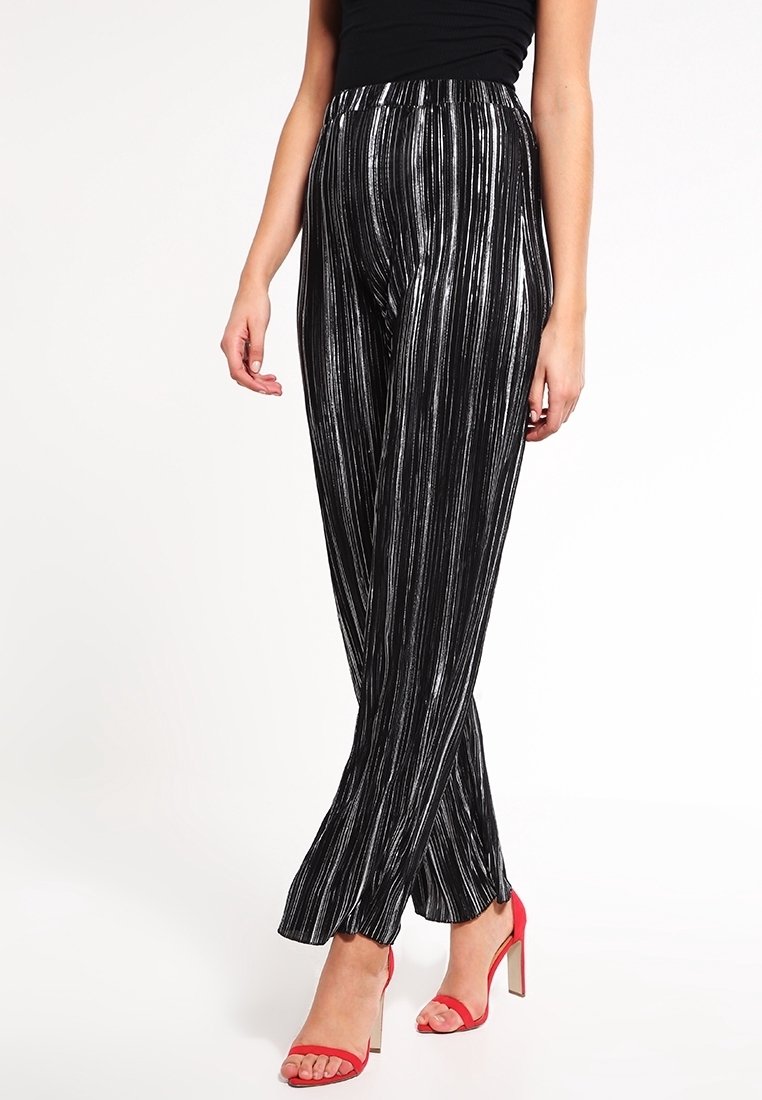 Femme portant un pantalon noir à jambes larges avec des rayures argentées verticales et des sandales rouges à talons hauts, debout sur un fond blanc.
