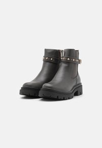 Bottes noires en cuir jusqu'à la cheville avec une semelle épaisse, fermeture éclair sur le côté, et une sangle décorative avec des clous en or. Finition texturée, conception robuste.