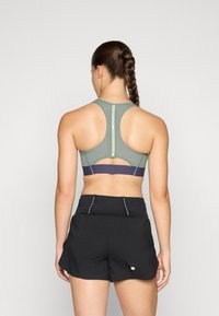 Zelena sportska grudnjak s dizajnom racerback, sa tamnoplavim pojasom na dnu. Crne trkačke šortse sa prednjim džepovima i reflektirajućim logotipom.