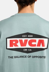 Light blue cotton t-shirt featuring a large graphic on the back. The design includes a red and black RVCA logo with the text "THE BALANCE OF OPPOSITES."