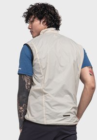 Leichter beiger Weste mit hohem Kragen, aus glattem Synthetikmaterial. Mit kleinem Logo und kontrastierenden Borten entlang der Armausschnitte.