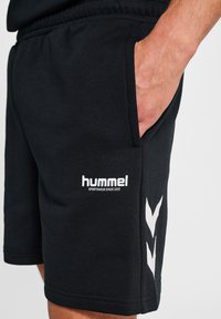 Man in zwarte Hummel sportshorts met een hand in de zijzak, witte logo en chevron ontwerp op de dij tegen een witte achtergrond.