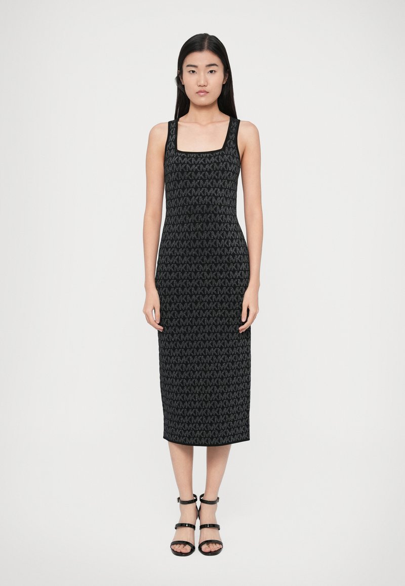 Čierne šaty strednej dĺžky so štvorcovým výstrihom a textúrovaným vzorom z opakujúcich sa log "MK". Dopĺňajú ich čierne sandále s remienkami.