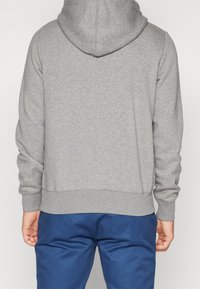 Felpa grigia con una texture morbida, dotata di un ampio cappuccio e polsini a costine. Indossata con pantaloni blu, che mostrano la vista posteriore.