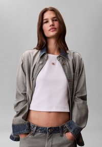 Ung kvinde med brunt hår bærer en lys denimjakke over en hvid cropped tanktop og matchende denim jeans mod en ensfarvet grå baggrund.