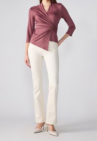 Lila kääritopp, jossa on kierre yksityiskohtana vyötäröllä, kolmasosapituudet hihat, yhdistetty valkoisiin kapeisiin housuihin ja vaaleanbeigeihin korkeisiin korkoihin.