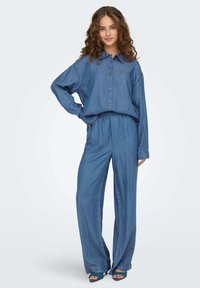 Džínový outfit zahrnující volnou košili na knoflíky s límcem a širokými kalhotami. Texturovaná modrá látka s elastickým pasem.