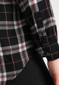 Sleeve of black, white, and red plaid shirt with buttoned cuff over black denim fabric and exposed hand.