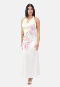 Femme avec les cheveux raides et foncés portant une robe maxi sans manches jaune avec des motifs floraux roses et des sandales à talons blancs.