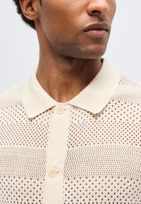 Άνδρας που φοράει πλεκτό πόλο πουκάμισο σε κρεμ χρώμα, με κοντά μανίκια και κουμπιά, με διάτρητο μοτίβο, κοιτάζοντας πλάγια μπροστά σε απλό φόντο.