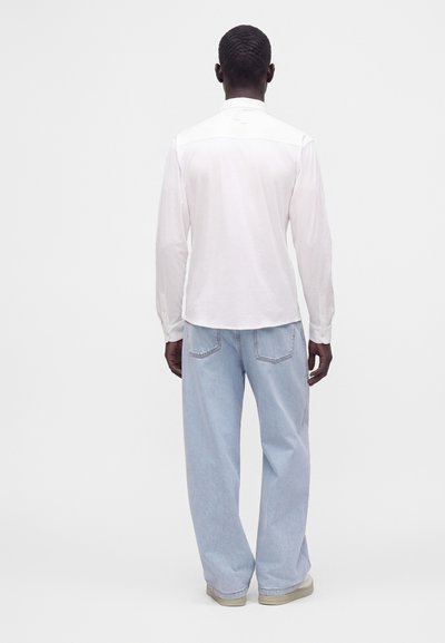 Homme aux cheveux courts portant une chemise blanche à manches longues et un jean bleu clair à jambes larges, debout de dos sur un fond uni.