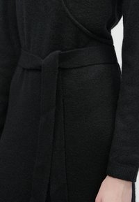 Svart strikket omslagskjole med en knyttet midjebelte og lange ermer, båret av en person med synlig høyre hånd og arm.