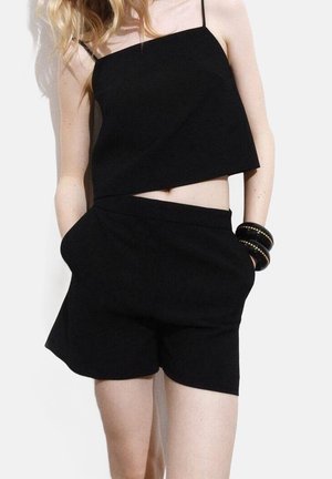 Femme portant un crop top noir sans manches et un short taille haute assorti, les mains dans les poches et des bracelets rigides noirs au poignet.