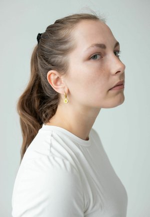 Young woman with brown hair in a ponytail wearing white shirt and gold hoop earrings with small daisy charms, looking sideways against plain background.