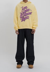 Gelbe Baumwoll-Hoodie mit fettem lila Text "Low Lights & Summer Nights", kombiniert mit dunklen Jeans und beigem Schuhwerk.