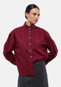 Chemise bourgogne à boutons, avec un col montant, un devant plissé et des manches bouffantes volumineuses, fabriquée à partir d'un tissu lisse. Boutons de haute qualité présents.