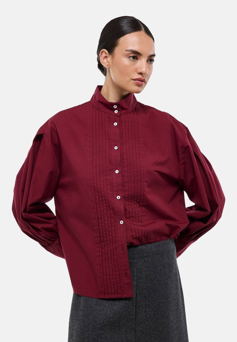Chemise bourgogne à boutons, avec un col montant, un devant plissé et des manches bouffantes volumineuses, fabriquée à partir d'un tissu lisse. Boutons de haute qualité présents.