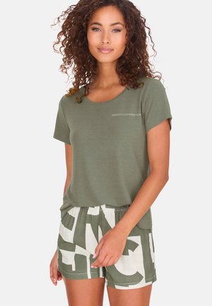 Frau mit lockigem Haar, die ein olivgrünes "French Connection"-T-Shirt und abstrakt gemusterte Shorts trägt, steht mit einer Hand entspannt an der Seite.