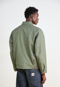 Grön, lätt jacka med krage, ryggparti och två knappar vid midjan. Modellen bär mörka denimbyxor med en logotypapplikation.