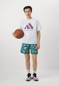 Hvid t-shirt med pink og lilla grafisk logo, teal shorts med hvide leopardpletter, hvide sokker og sorte og røde sneakers. Holder en basketball.