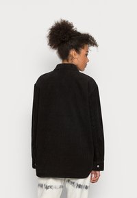 Casaco oversized de cabedal preto com colarinho, mangas longas e punhos com botão. O tecido texturizado apresenta riscas verticais.