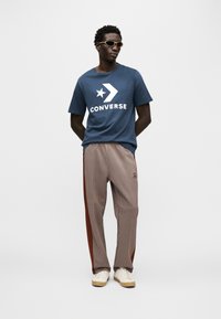 Μπλε βαμβακερό μπλουζάκι με λευκό λογότυπο "CONVERSE", συνδυασμένο με καφέ καρό παντελόνι με ρίγες στο πλάι και λευκά αθλητικά παπούτσια.