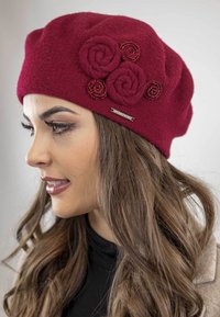 Berretto in lana color marrone porpora con tre rose in feltro e accenti a spirale decorativi, completato da un'etichetta metallica sul lato.