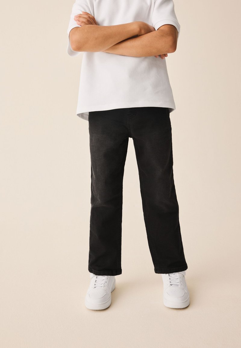 Kind steht mit verschränkten Armen, trägt ein weißes kurzärmeliges Hemd, schwarze Hose und weiße Turnschuhe vor neutralem Hintergrund.